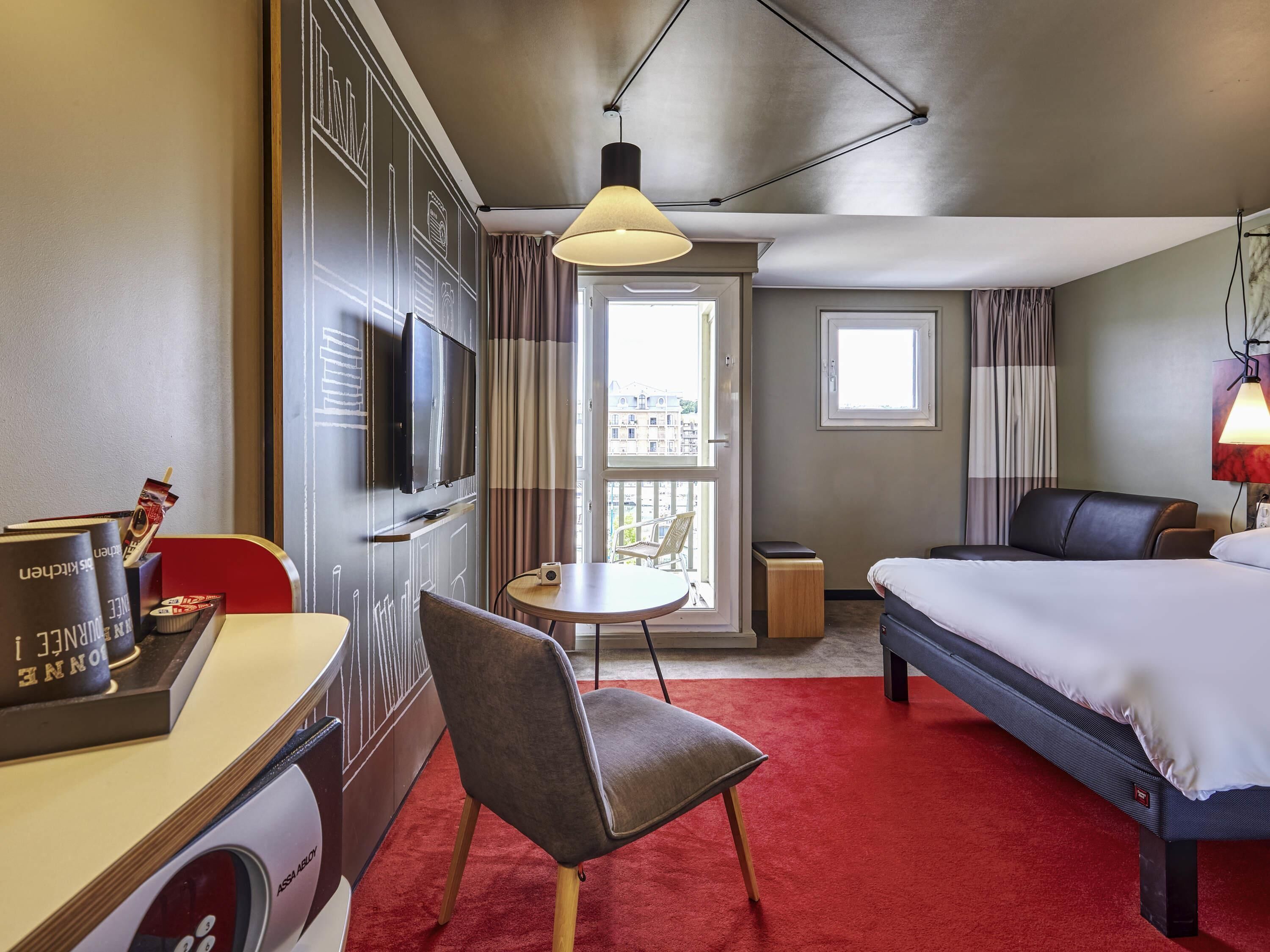A hotel room with a bed, a desk with a chair, a television, a small table, and a balcony with a view of buildings.