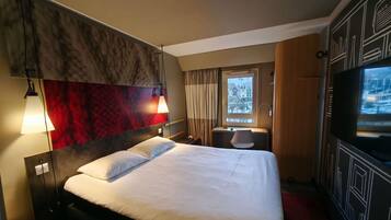 Habitación de hotel con cama, escritorio, silla, televisor y ventana con vista a la ciudad.