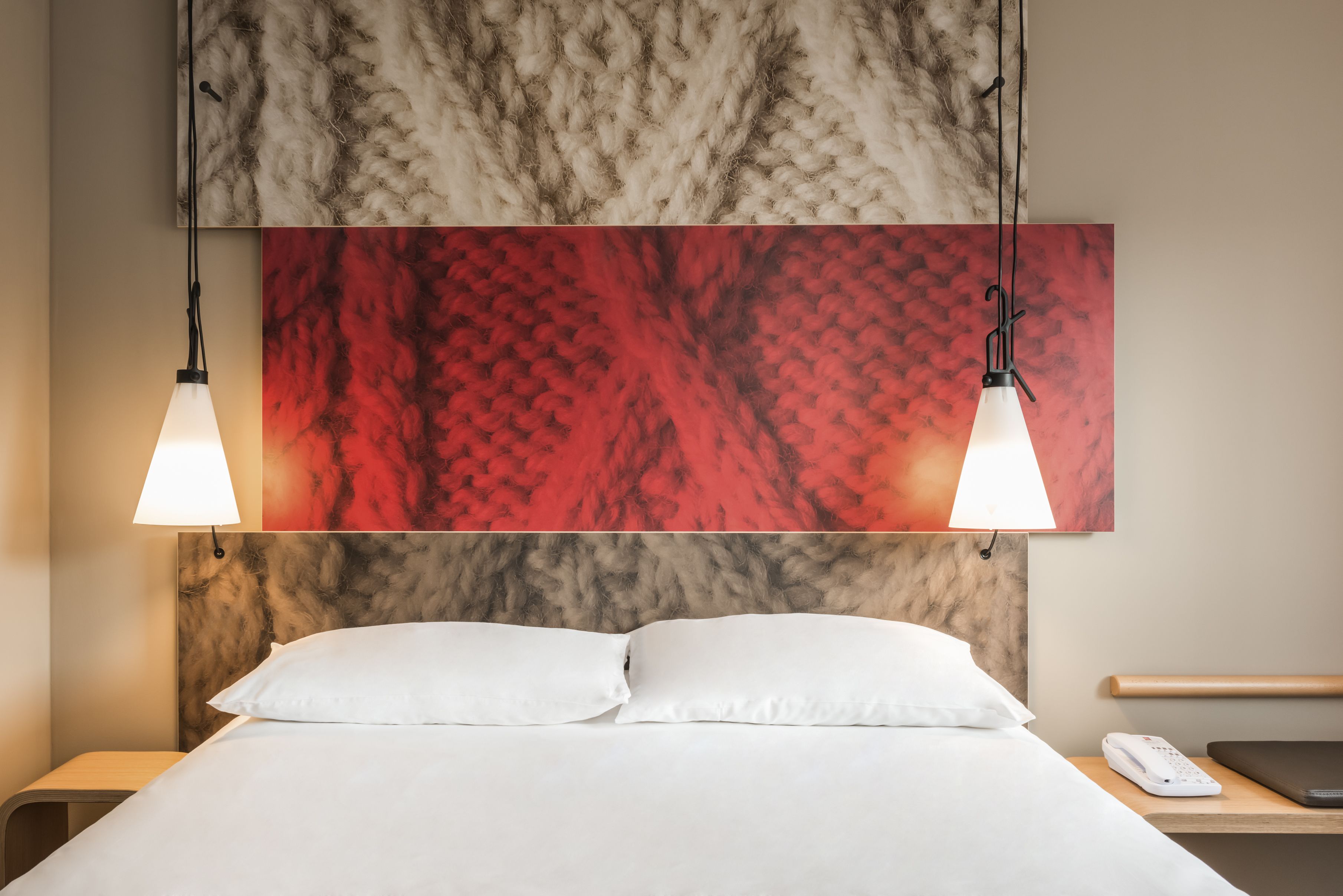 Una habitación de hotel con una cama, dos lámparas de noche, una mesita con teléfono y un cuadro abstracto grande sobre la cama.