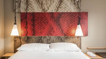 Una habitación de hotel con una cama, dos lámparas de noche, una mesita con teléfono y un cuadro abstracto grande sobre la cama.