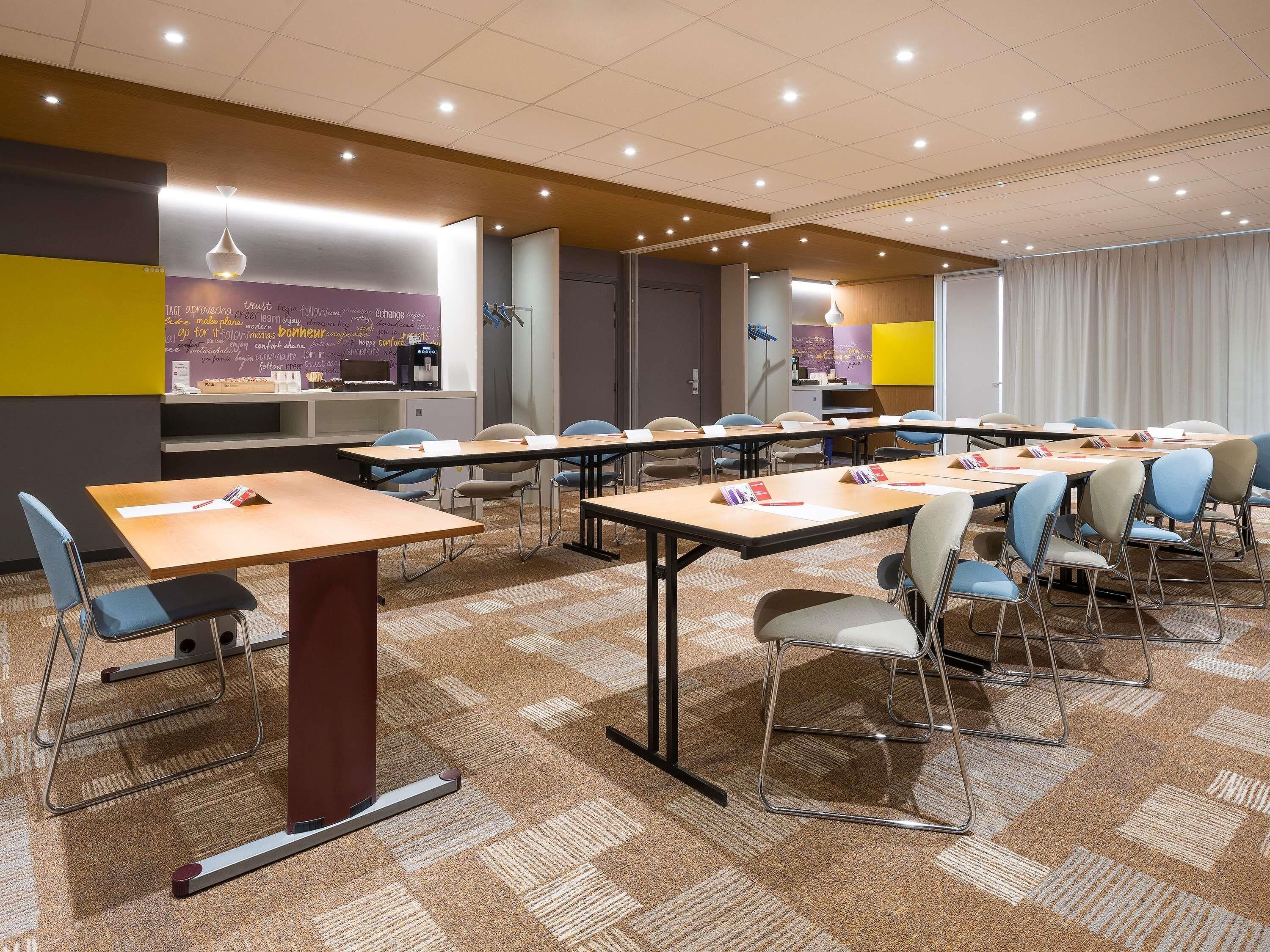 A conference room with multiple tables and chairs, a kitchenette, and a view of the cityscape through the windows.