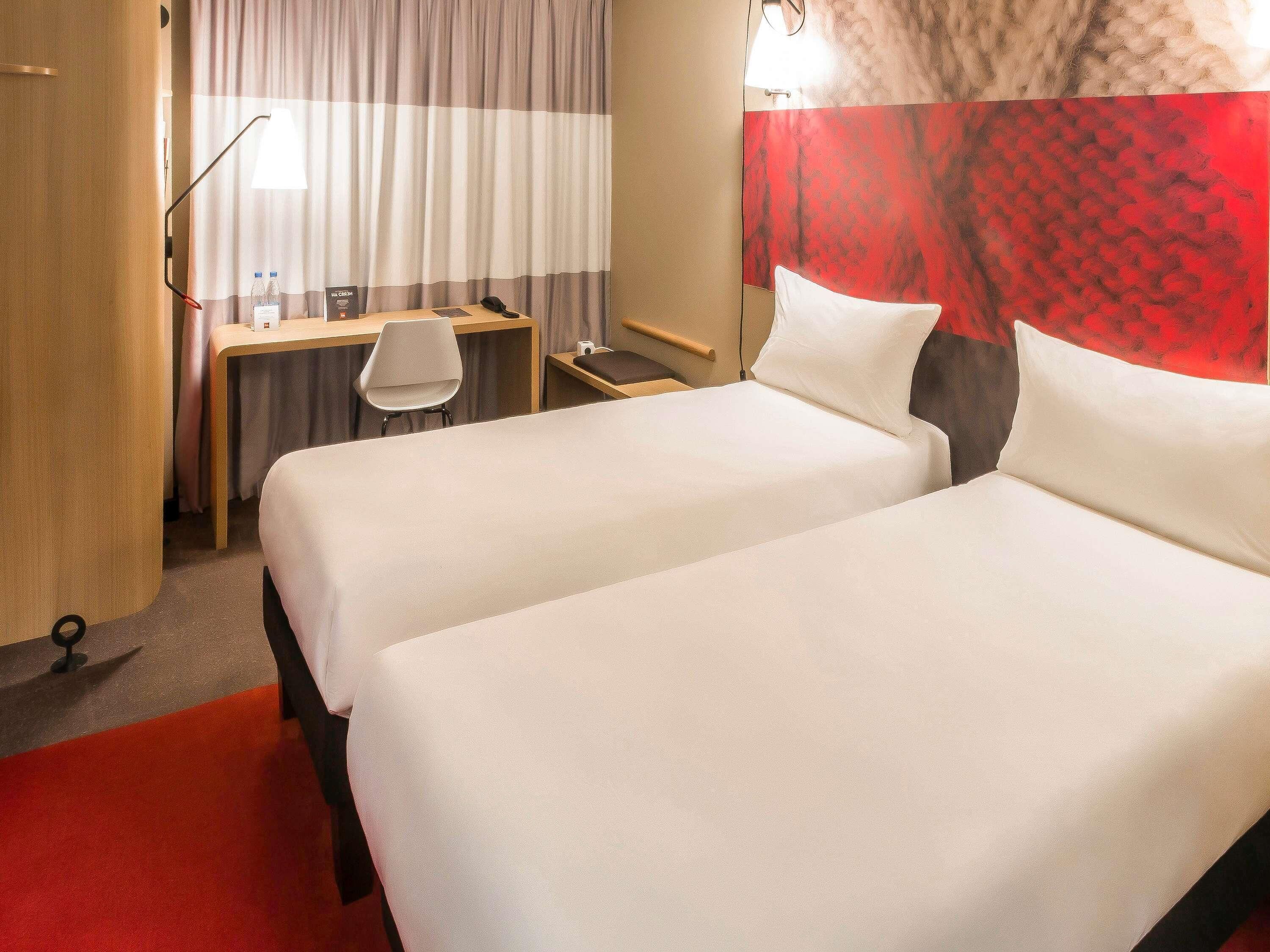Una camera d'albergo con due letti, una scrivania, una sedia, un telefono e una parete con un motivo rosso decorativo.