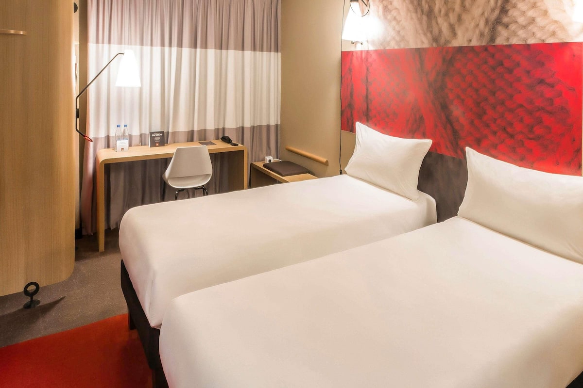 A hotel room with two beds, a desk, a chair, a telephone, and a wall with a red patterned design.
