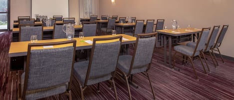 A conference room with multiple tables and chairs, a projector screen, and a carpeted floor.
