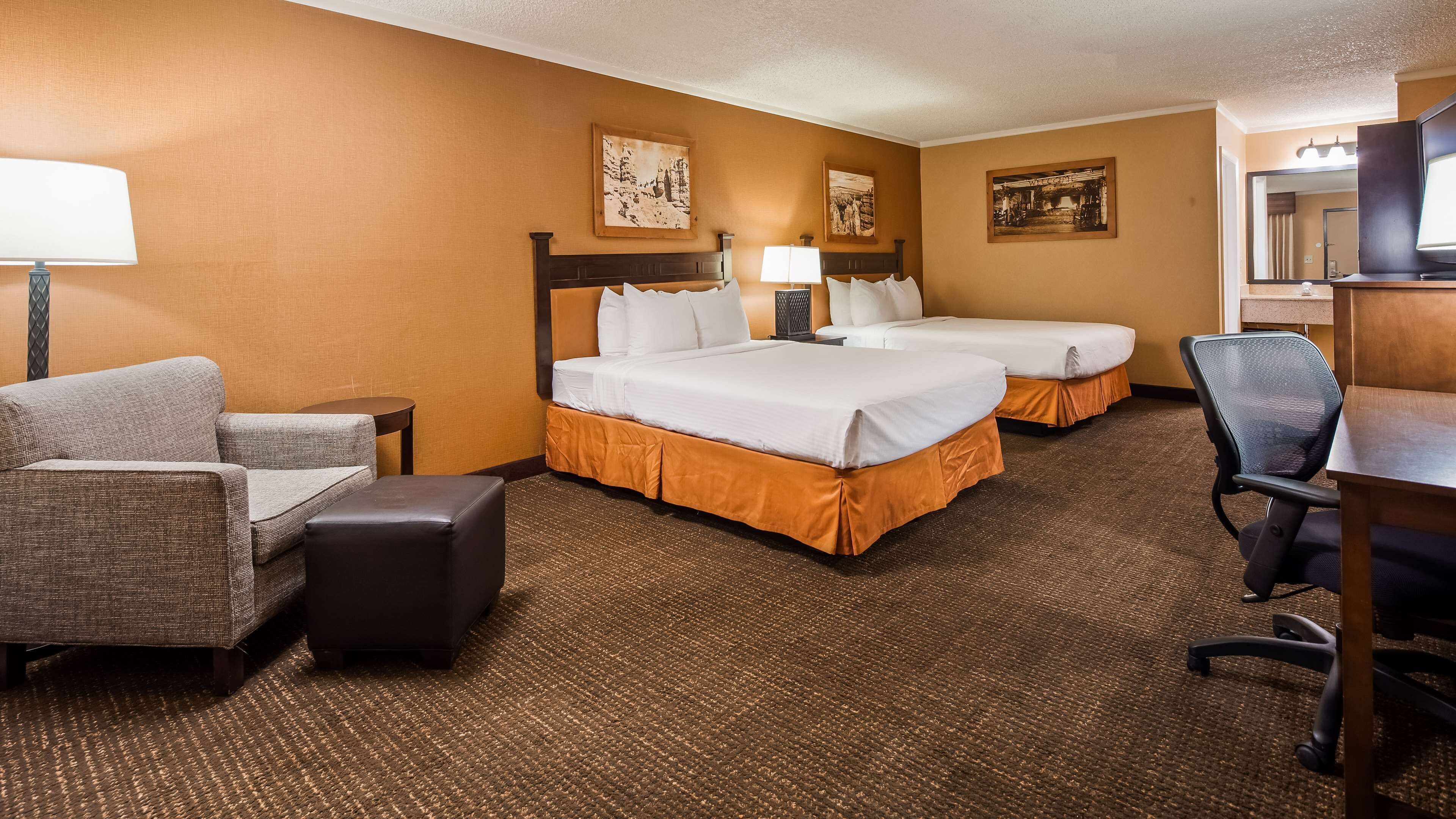 A hotel room with two beds, a desk, a chair, a lamp, and two wall-mounted paintings.