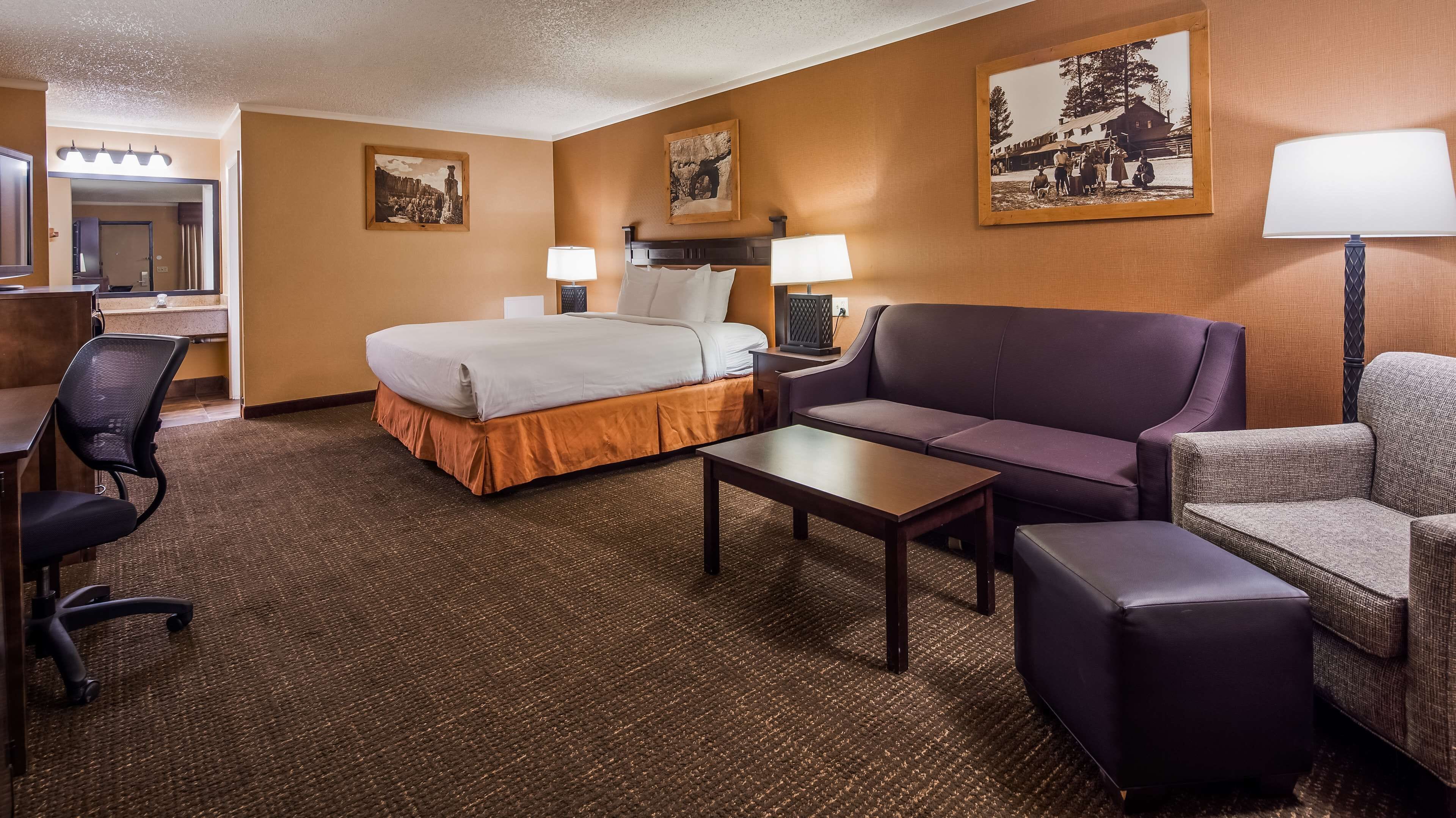 A hotel room with a bed, two bedside tables, a sofa, a chair, a small table, and a desk with a chair.