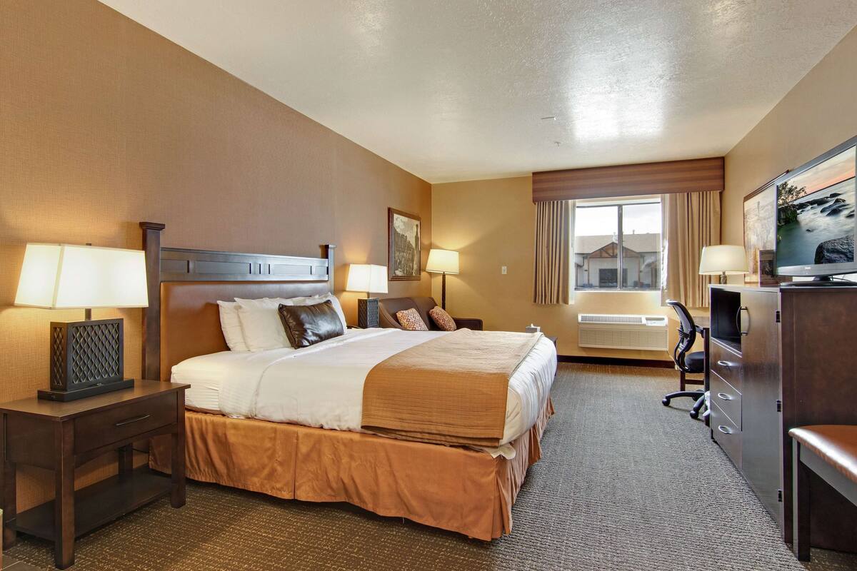 A hotel room with a large bed, two bedside tables with lamps, a desk with a chair, a television, and a window with curtains.