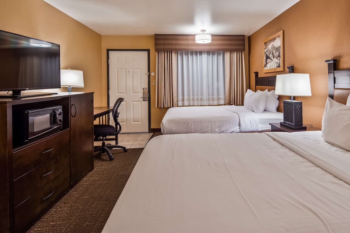 A hotel room with a bed, a desk with a TV, a microwave, a chair, a nightstand with a lamp, and a door.