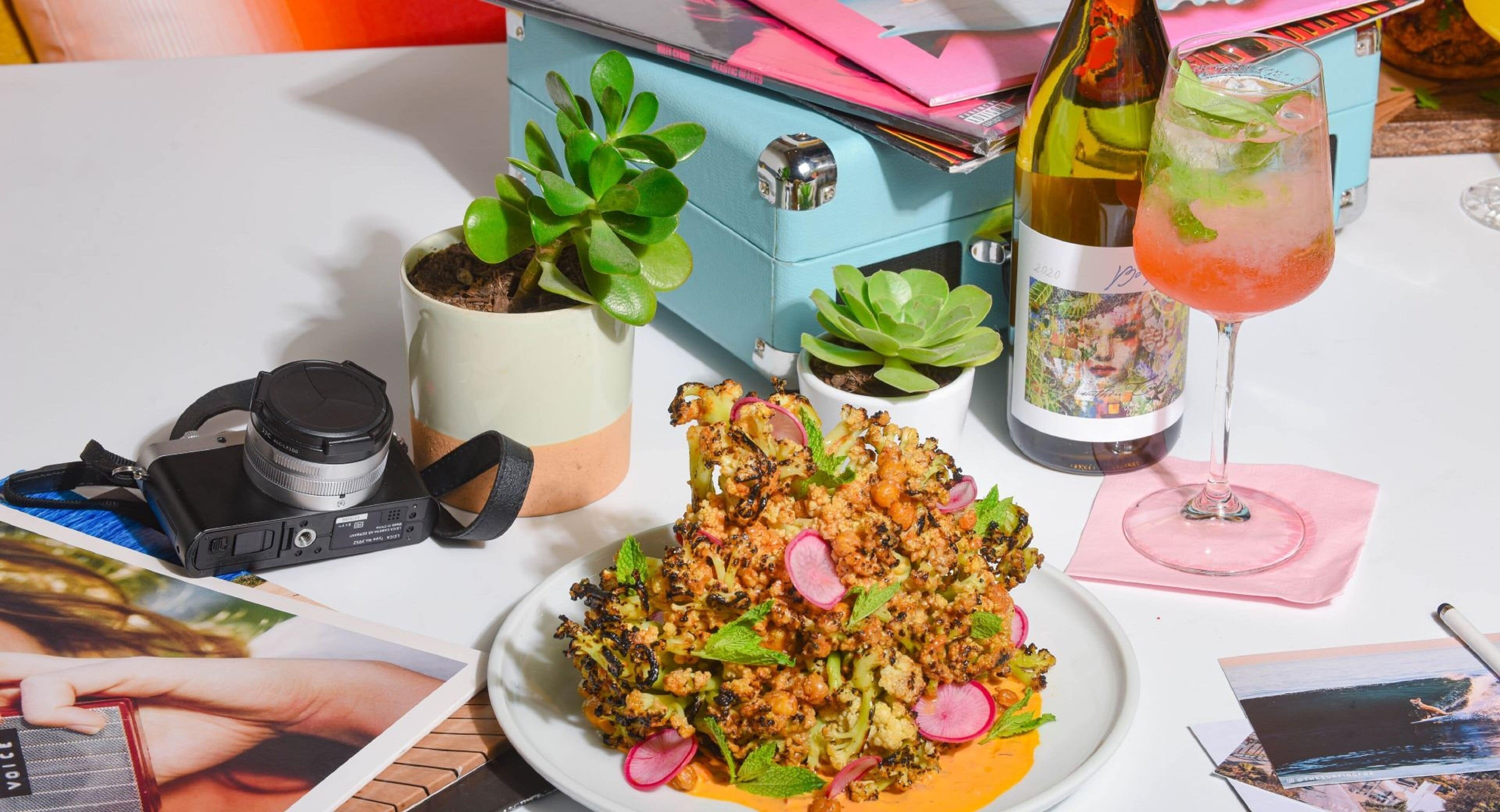 A table with a plate of food, a camera, a wine bottle, and a drink.