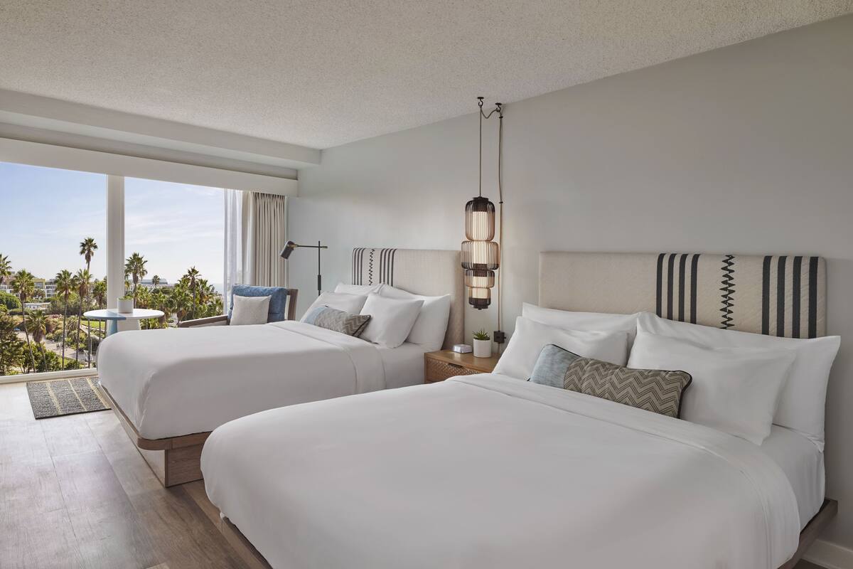 A hotel room with two beds, a large window with a view, a bedside table with a lamp, and a view of the outdoors.