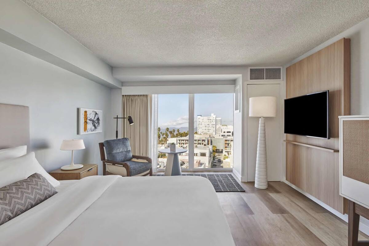 A hotel room with a large bed, a desk with a lamp, a chair, a TV, and a view of the city through large windows.