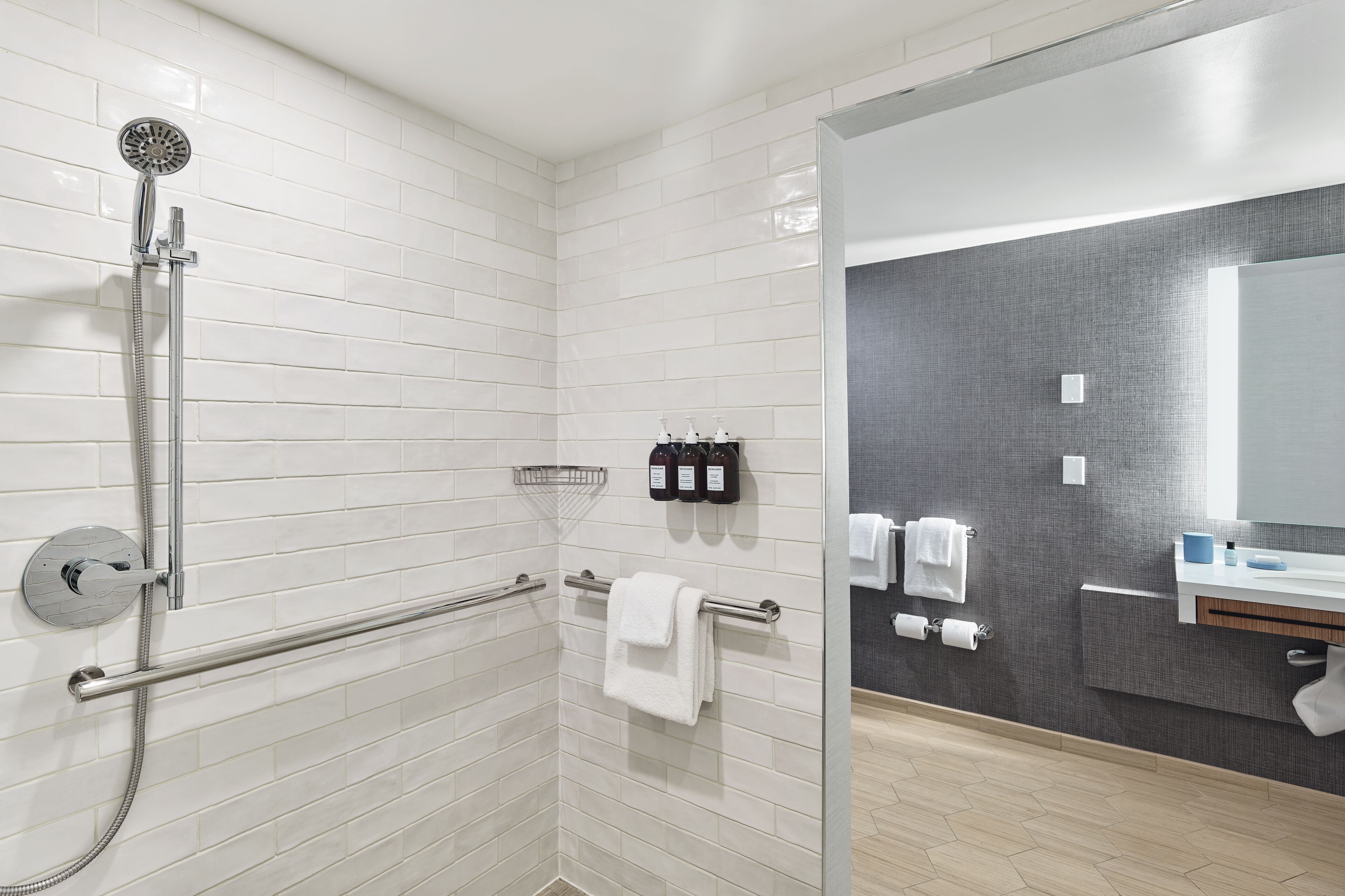 Un baño moderno con paredes de azulejos blancos, cabina de ducha con mampara de vidrio, barra de apoyo, toallero y tocador con espejo.