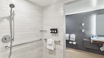 Un baño moderno con paredes de azulejos blancos, cabina de ducha con mampara de vidrio, barra de apoyo, toallero y tocador con espejo.