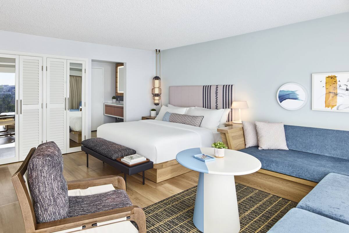 A hotel room with a bed, a sofa, a chair, a small table, and a desk with a lamp. There is a window with a view of the city.