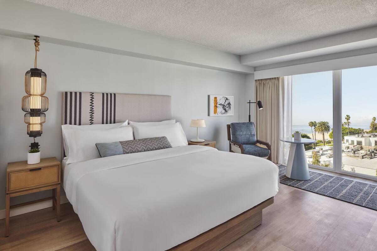 A hotel room with a large bed, a bedside table with a lamp, a chair, and a view of the beach.