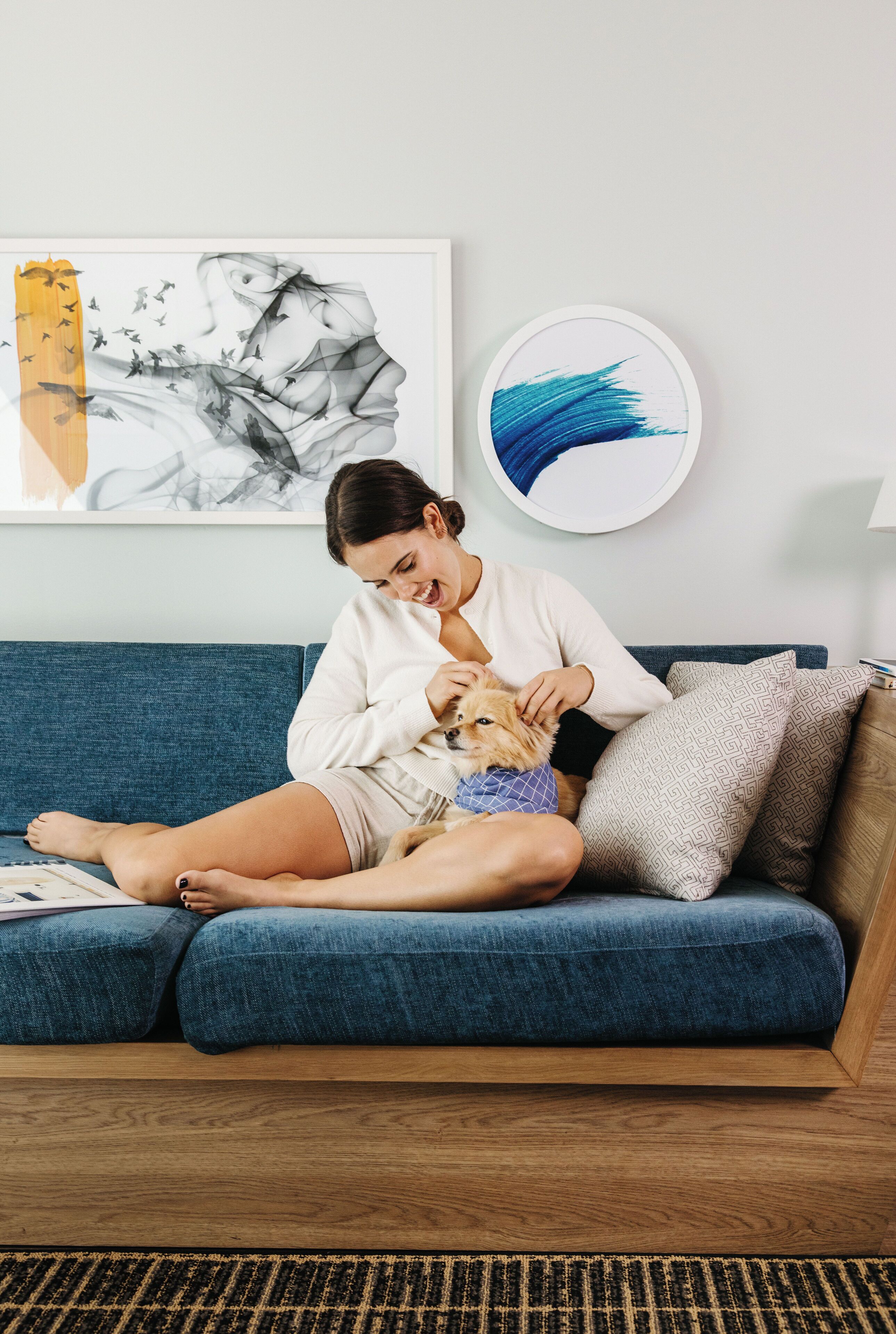 A person sitting on a blue sofa with a dog, surrounded by artwork on the wall.