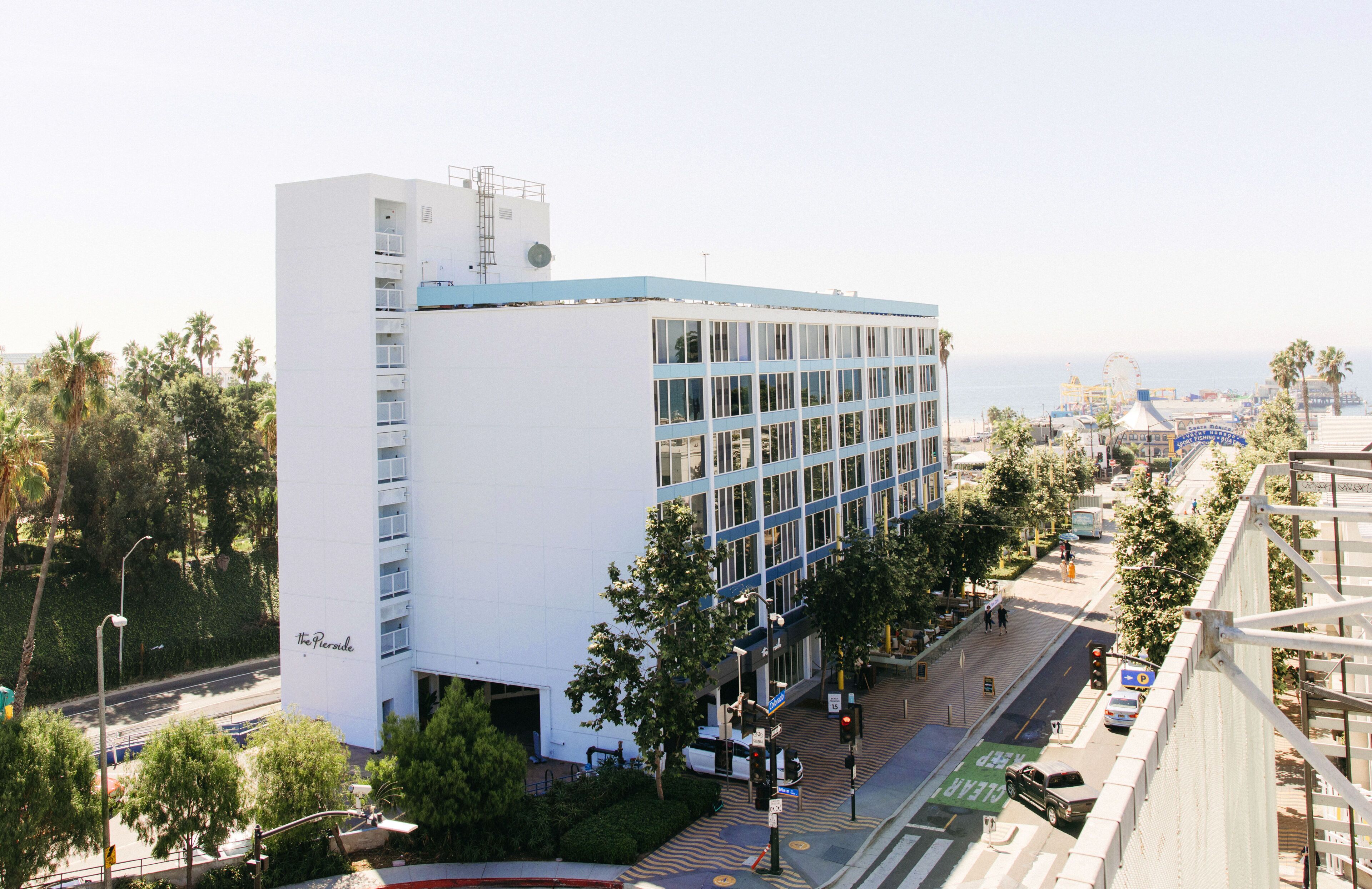 A modern hotel with a white facade and blue accents, located near a street with palm trees and a Ferris wheel in the background.