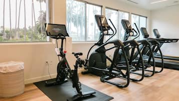 A gym with exercise bikes and treadmills, large windows with a view of palm trees, and a wooden floor.