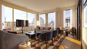 A modern living room with a sofa, chairs, a coffee table, and a TV. Large windows offer a city view.