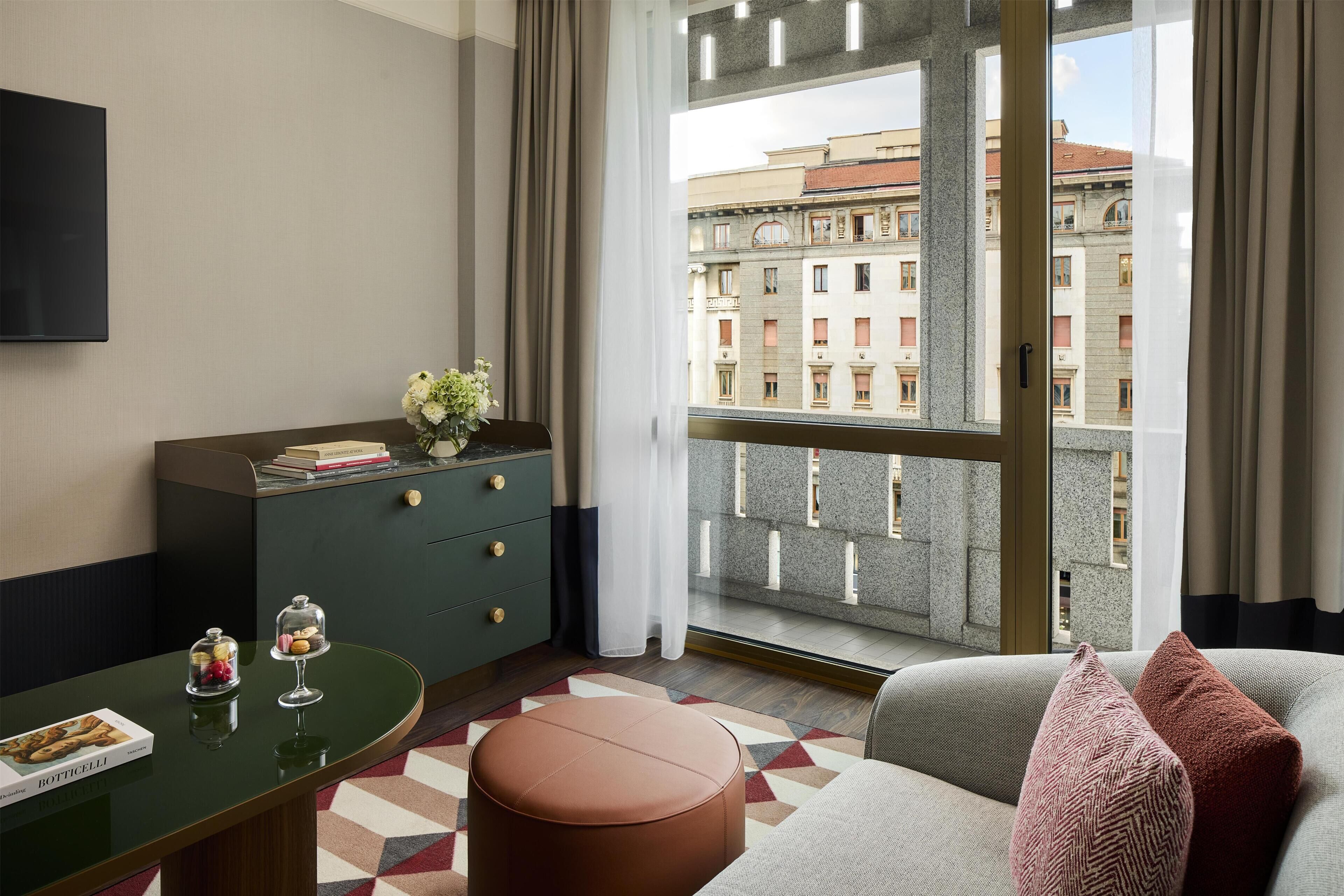 A modern hotel room with a large window, a sofa, a small table with a book, and a cabinet with decorative items.