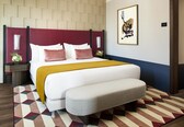 A modern hotel room with a large bed, a bedside table, a bench, a patterned rug, and a framed artwork on the wall.