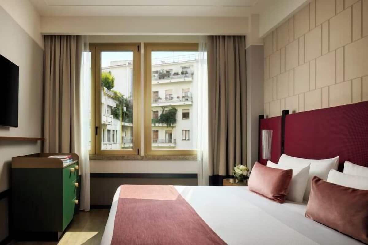 A hotel room with a bed, a dresser, a TV, and a window with curtains.