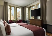 A hotel room with two beds, a TV, a desk with a chair, and large windows.