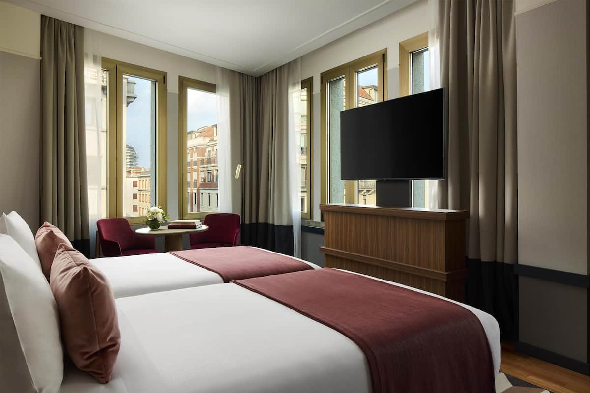 A hotel room with two beds, a TV, a desk with a chair, and large windows.
