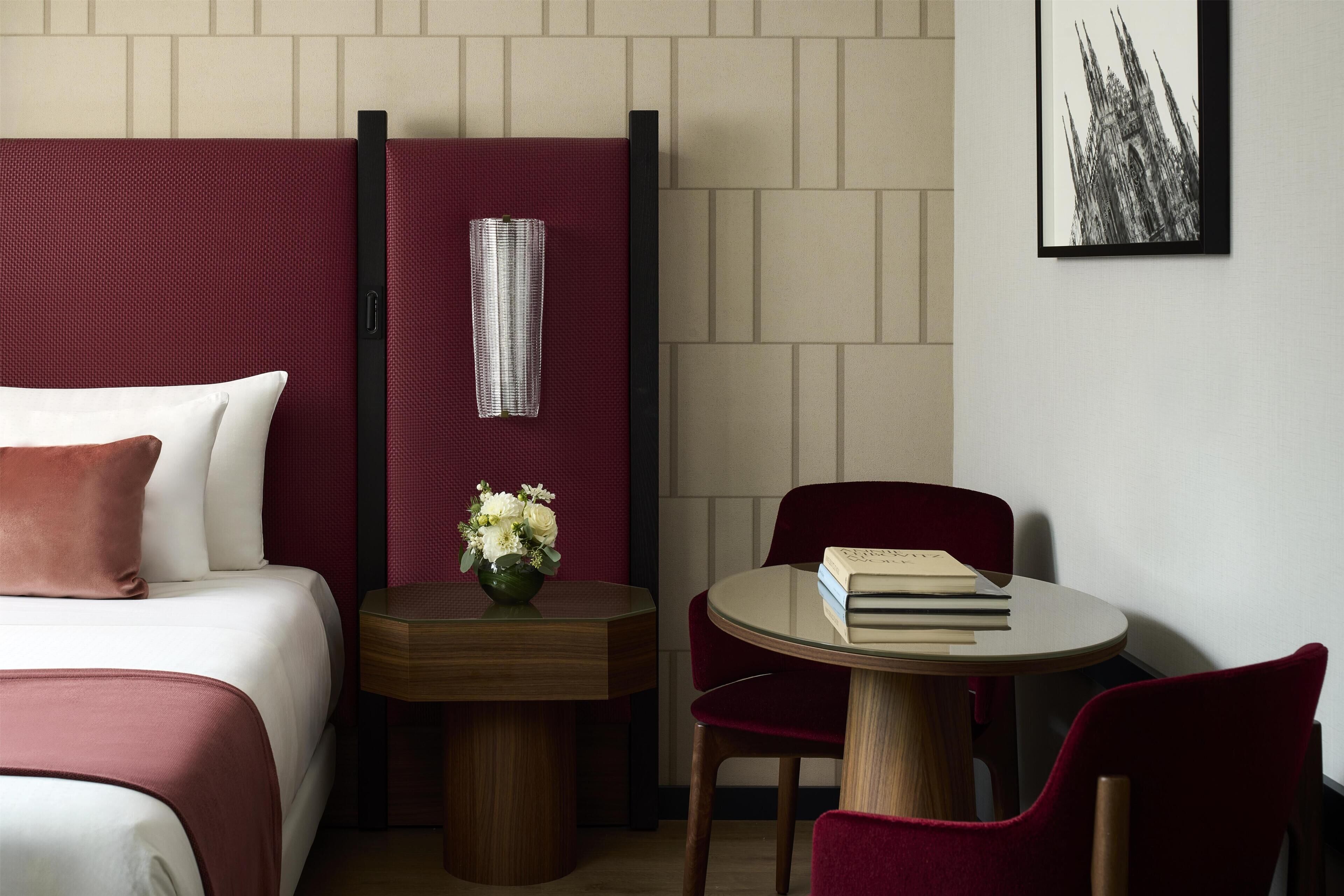 Una camera d'albergo con un letto, un tavolino con un vaso di fiori, un tavolo rotondo con sedie e un quadro appeso al muro.