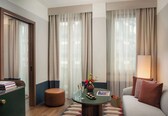 A hotel room with a sofa, a table with a vase of flowers, a lamp, a small round ottoman, and a bookshelf with books.