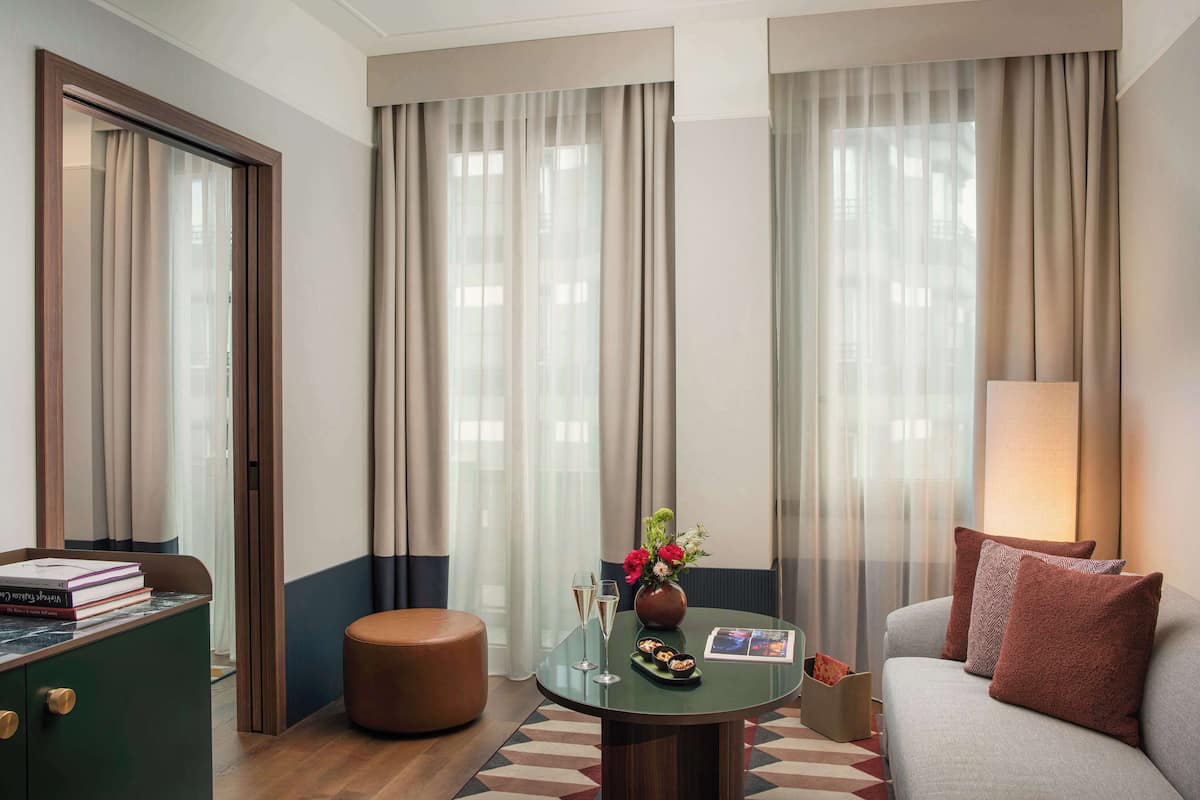 A hotel room with a sofa, a table with a vase of flowers, a lamp, a small round ottoman, and a bookshelf with books.