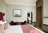 A hotel room with a bed, a desk, two chairs, a round table, a framed picture, a mirror, and a wooden door.