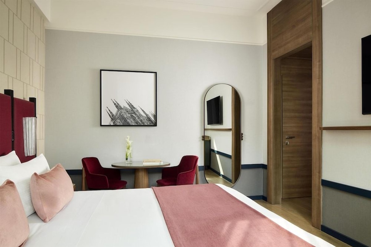 Een hotelkamer met een bed, een bureau, twee stoelen, een ronde tafel, een lijst, een spiegel en een houten deur.
