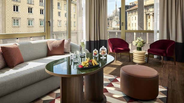 A modern hotel room with a sofa, a round table with fruit, two red chairs, and large windows with a city view.