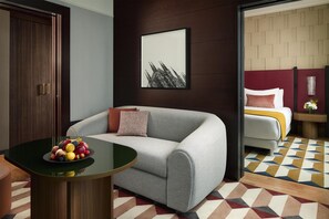 Une chambre d’hôtel moderne avec un canapé gris, une table ronde sur laquelle sont disposés des fruits, une œuvre d’art abstraite encadrée et un sol à motifs.
