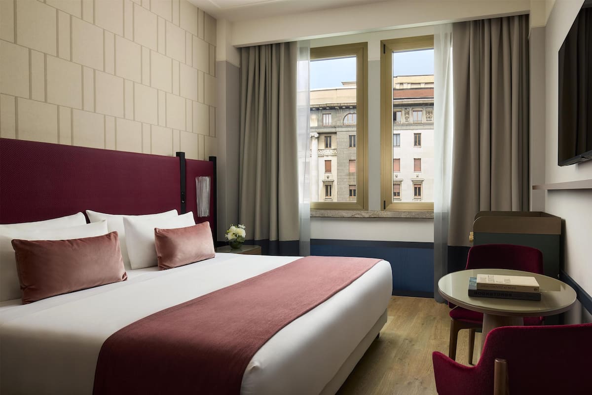 A hotel room with a large bed, a small table with a book, a chair, and a window with curtains.