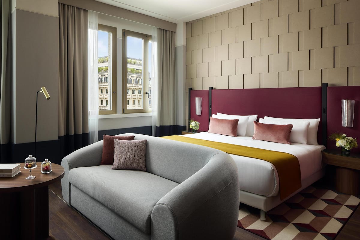 A hotel room with a bed, a sofa, a nightstand, a lamp, and a window with curtains.