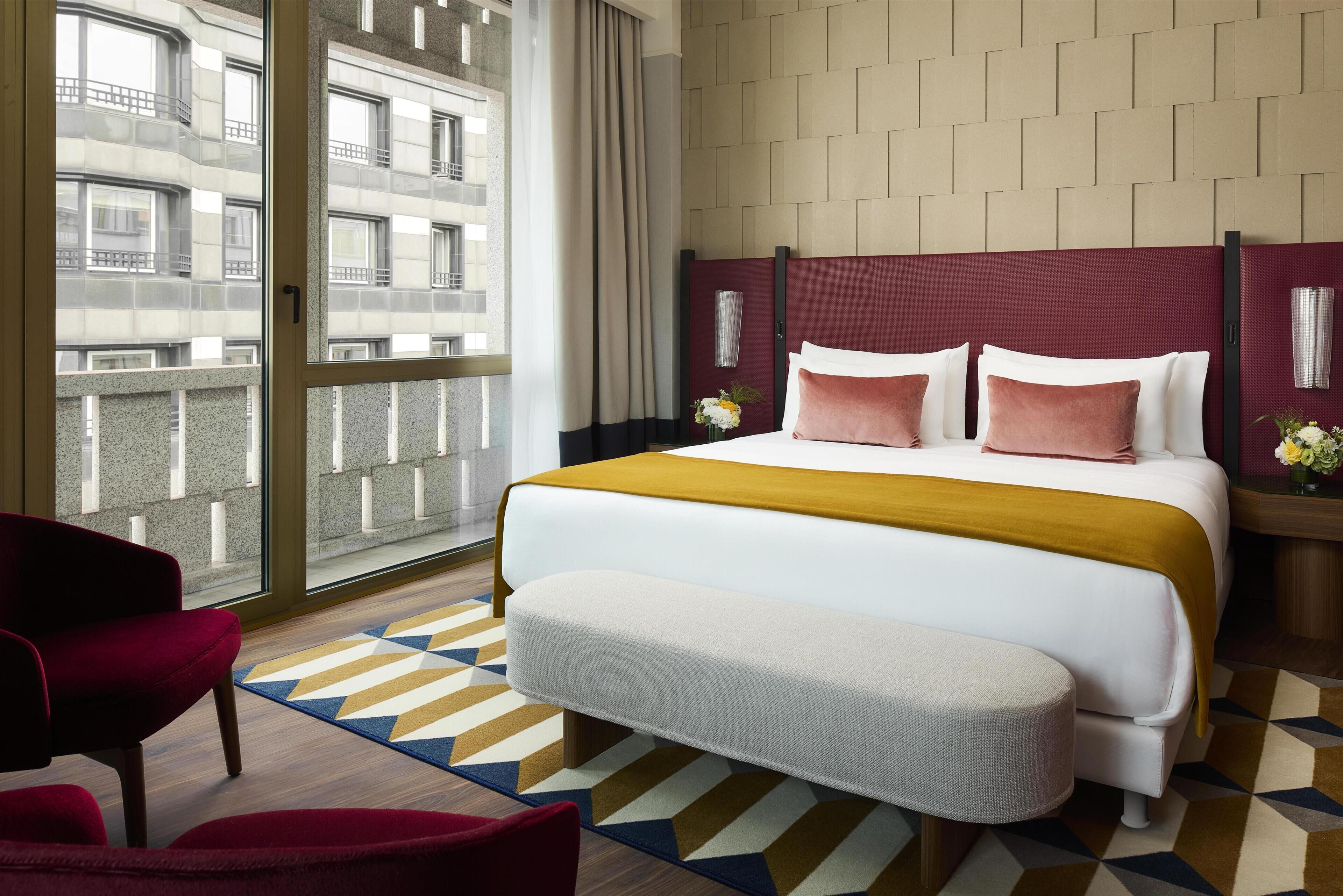 Habitación de hotel con una cama grande, una silla roja, un banco, un tapete estampado y vistas de la ciudad a través de un ventanal.