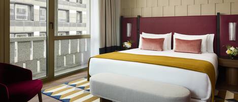 Habitación de hotel con una cama grande, una silla roja, un banco, un tapete estampado y vistas de la ciudad a través de un ventanal.