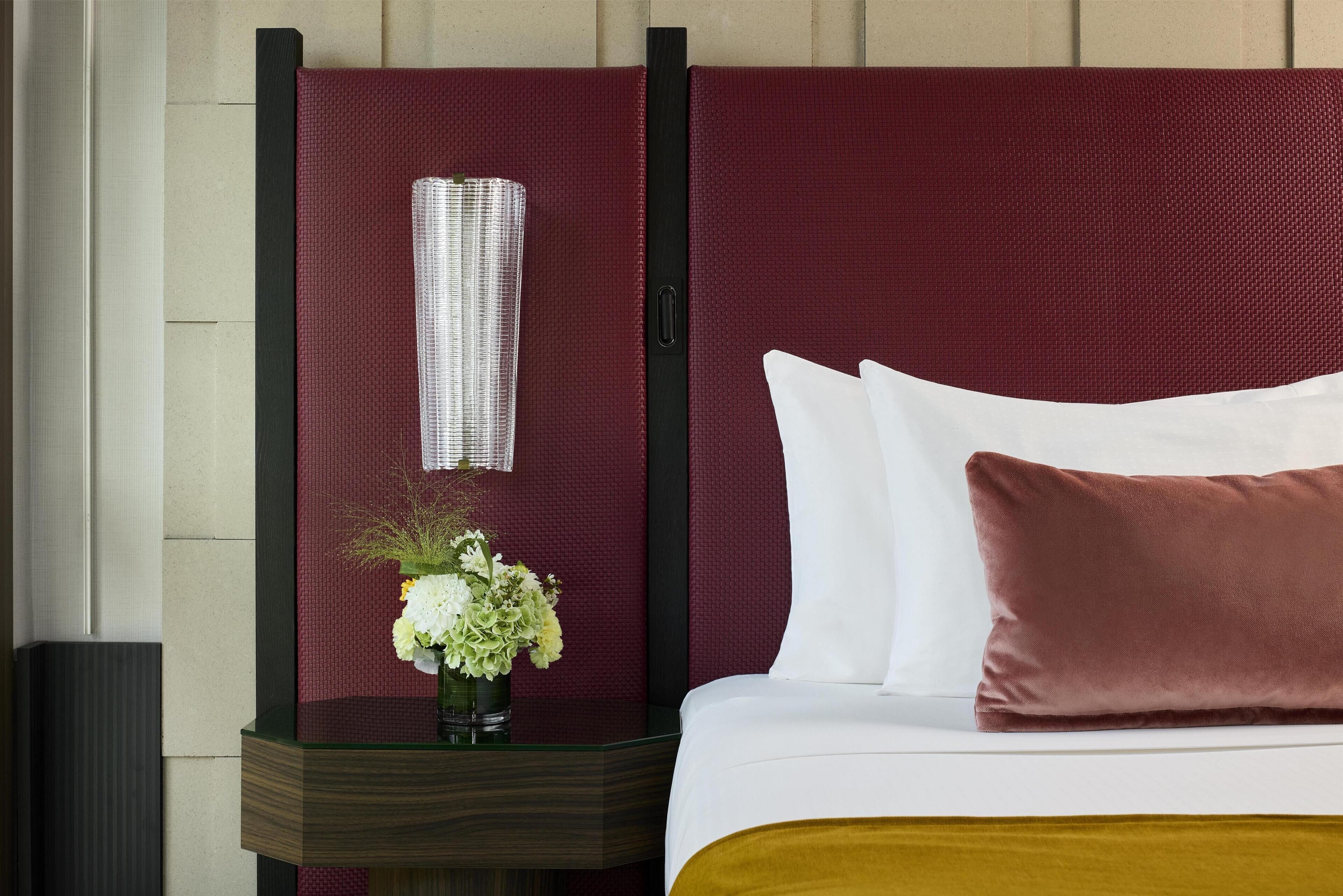 A hotel room with a bed, a nightstand, a vase with flowers, and a wall-mounted item.