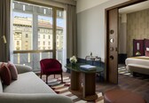 A hotel room with a sofa, a chair, a small table with a book, a vase with flowers, and a view of buildings through a large window.