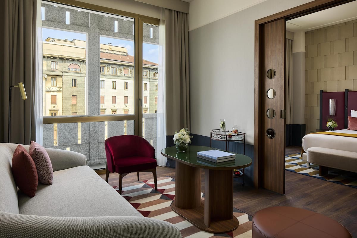 A hotel room with a sofa, a chair, a small table with a book, a vase with flowers, and a view of buildings through a large window.