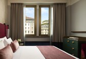 A hotel room with a bed, a nightstand, a dresser, and a window with a view of buildings.