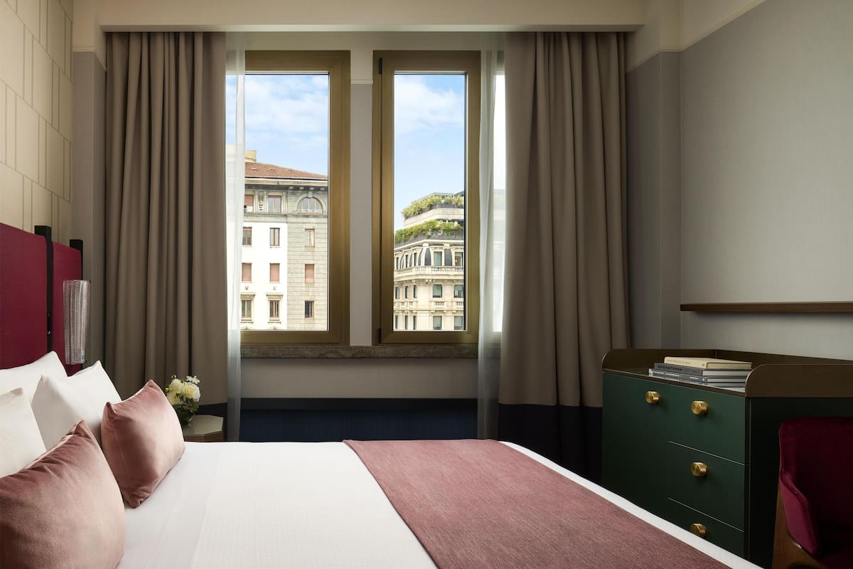 A hotel room with a bed, a nightstand, a dresser, and a window with a view of buildings.