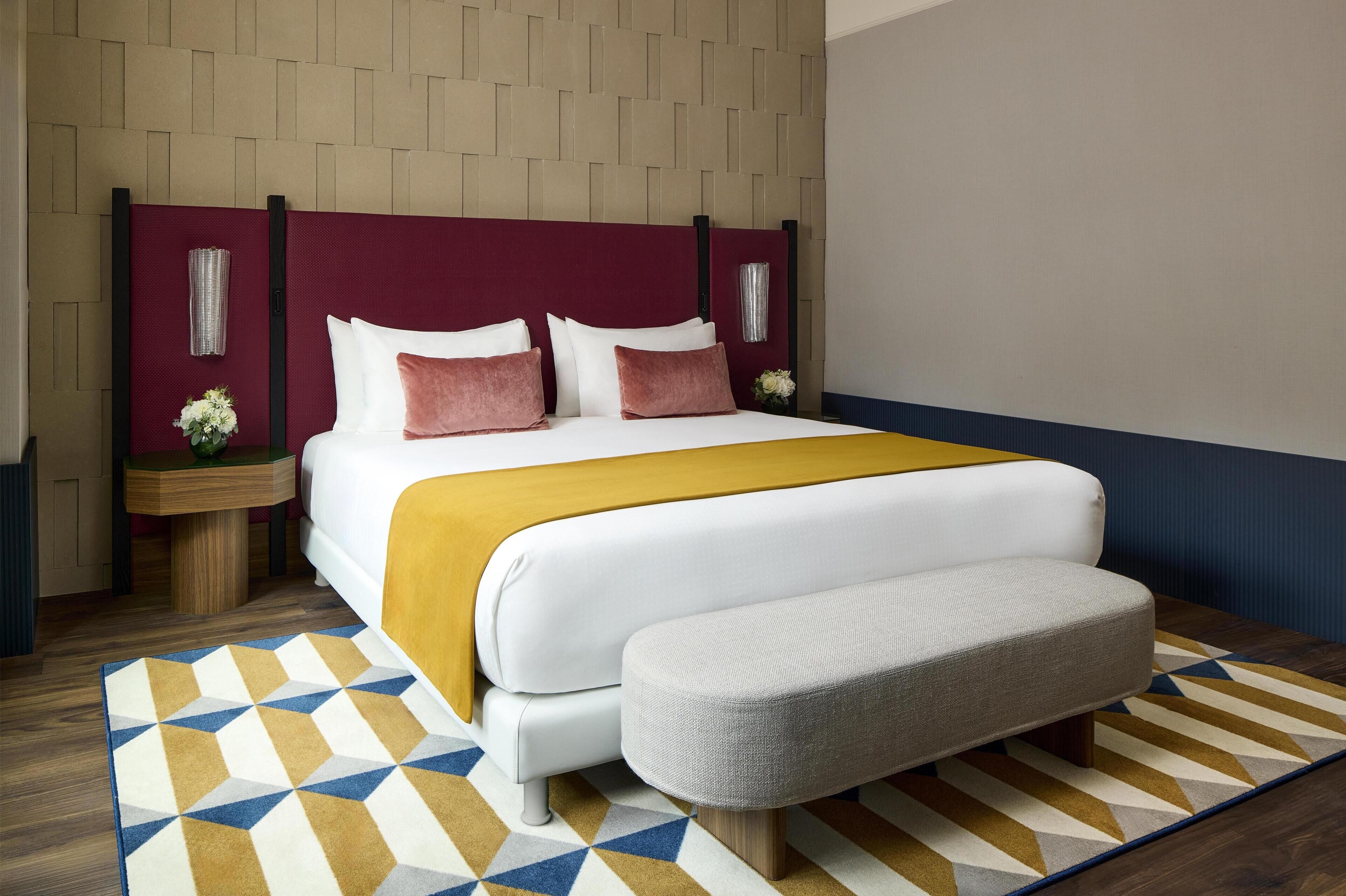 A modern hotel room with a large bed, a bench, a patterned rug, and a decorative wall panel.