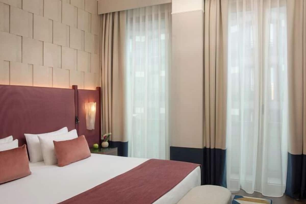 A hotel room with a large bed, a headboard with a light fixture, a nightstand with a vase, and sheer curtains.