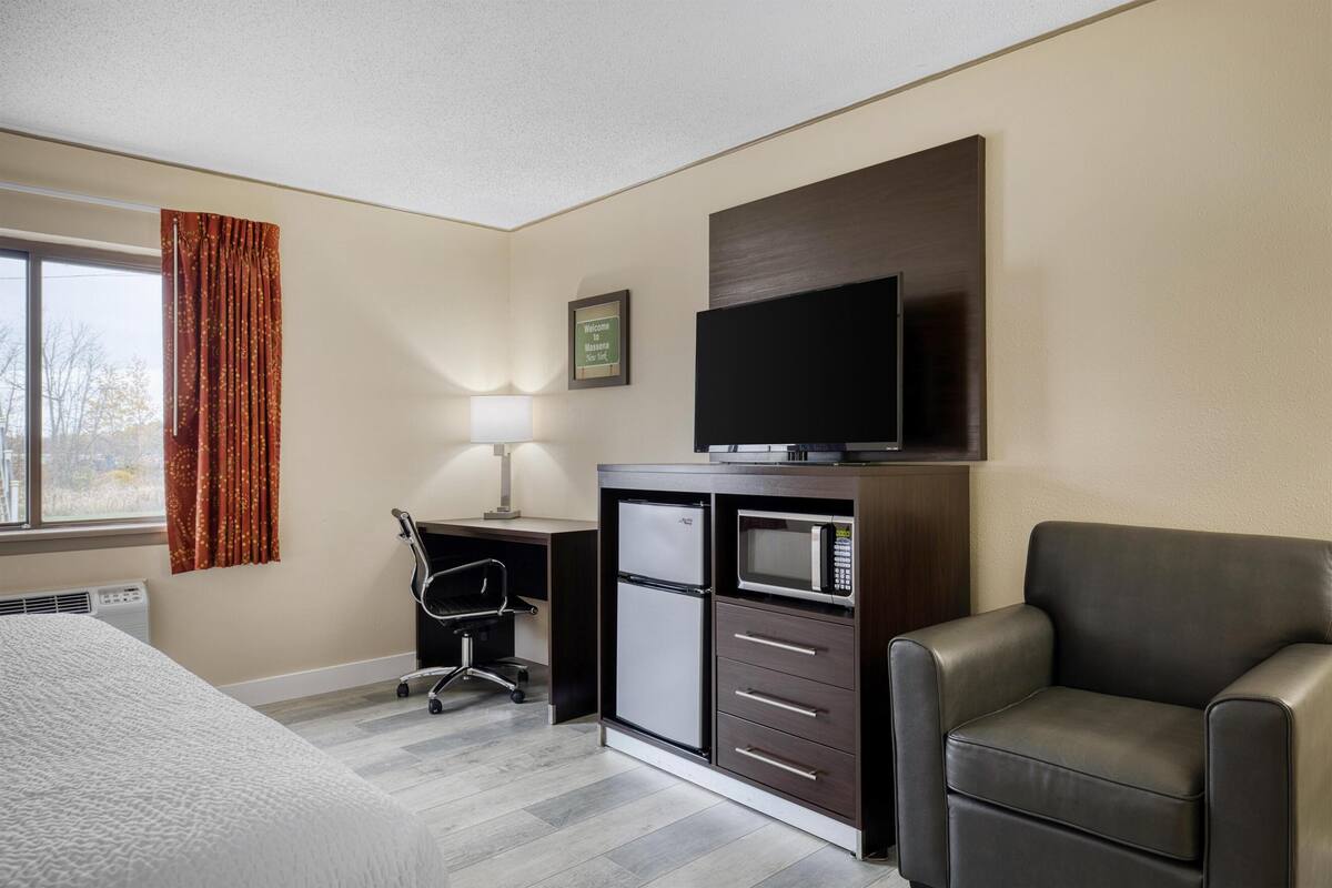Een hotelkamer met een bed, een bureau met stoel, een koelkast, een magnetron, een televisie en een leren fauteuil.