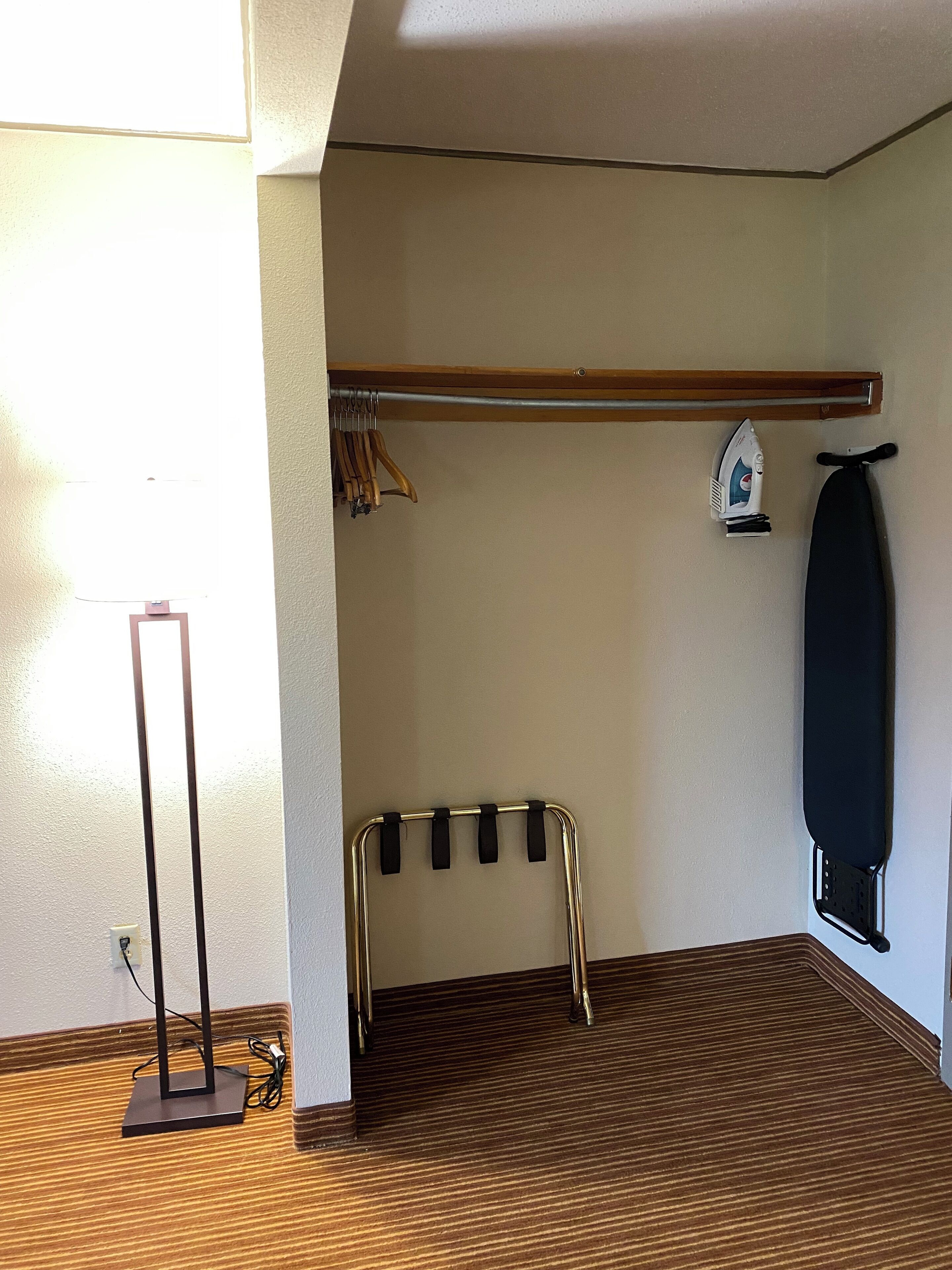 Quarto com piso de madeira, suporte de metal para cabides, tábua de passar roupa fixada na parede e uma luminária.
