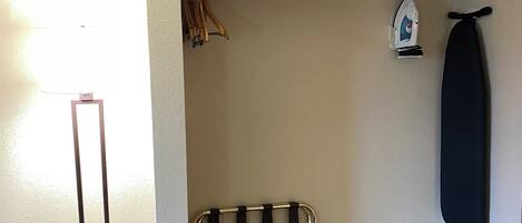 Quarto com piso de madeira, suporte de metal para cabides, tábua de passar roupa fixada na parede e uma luminária.