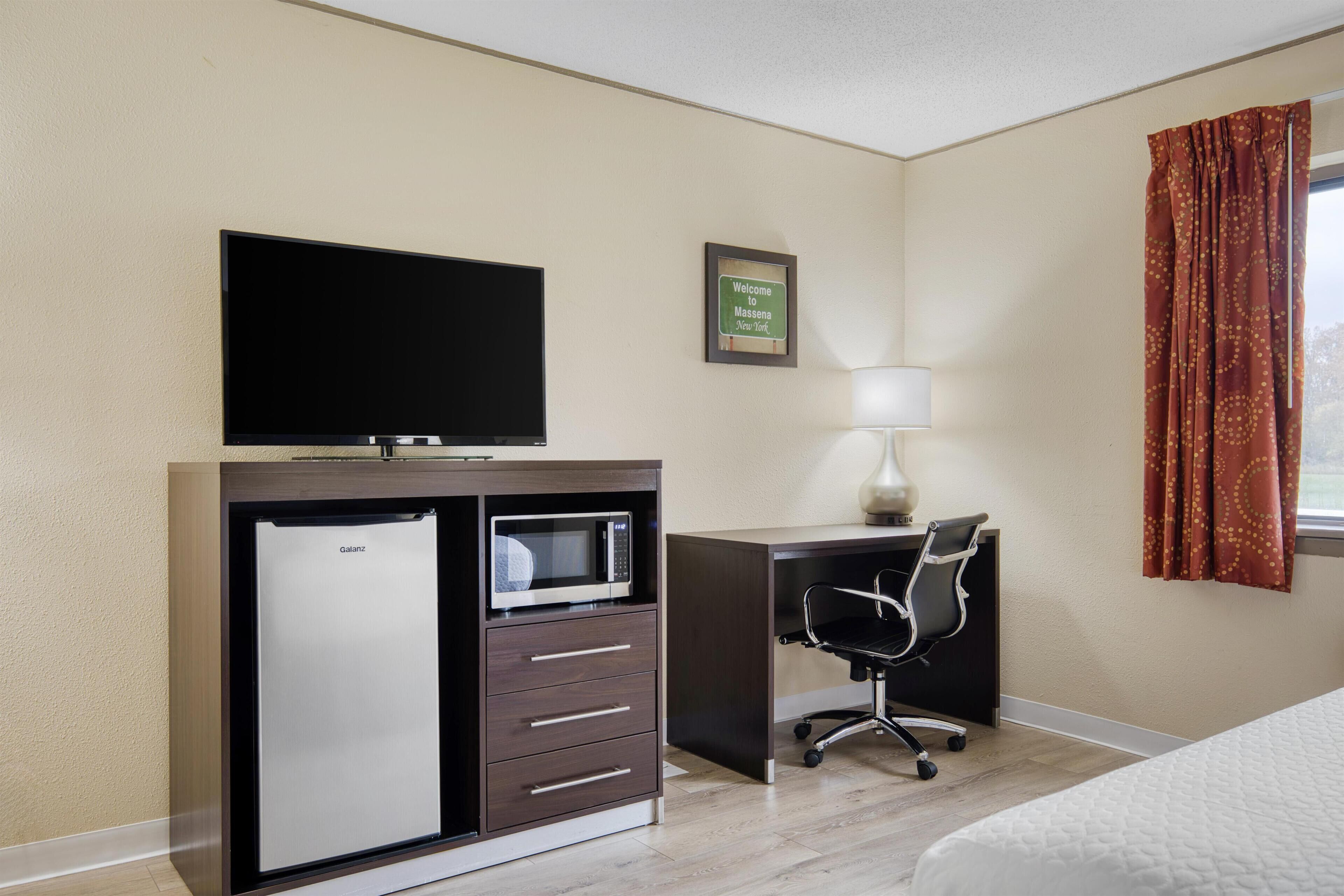 A hotel room with a flat-screen TV, a minibar, a desk with a chair, a lamp, and a bed.